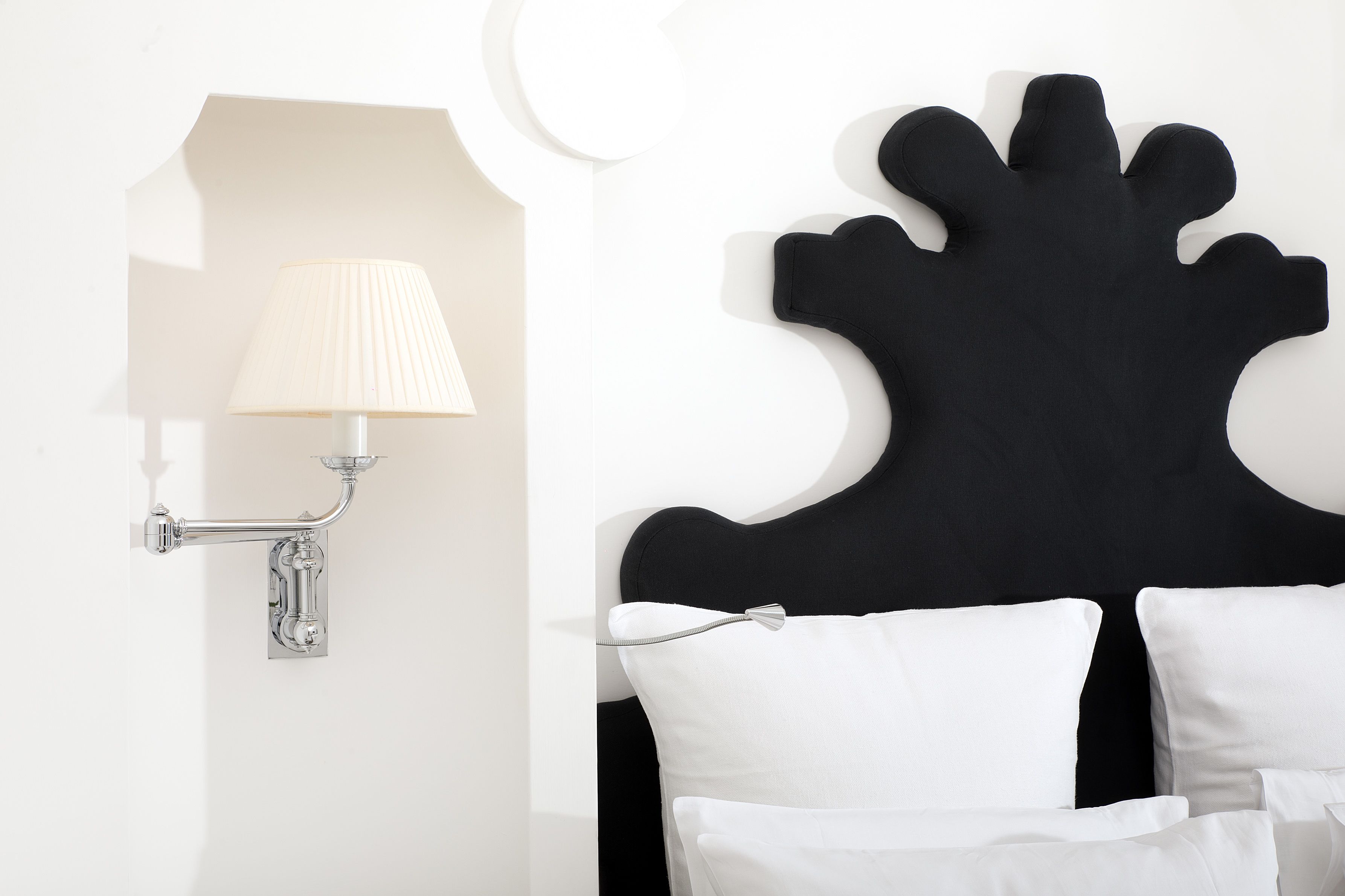 Una habitación moderna con cabecera negra, almohadas blancas y una lámpara fijada a la pared.