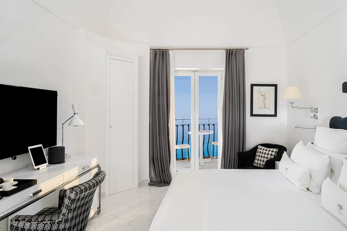 A hotel room with a bed, a desk with a TV, a chair, a balcony with a view of the sea, and a painting on the wall.