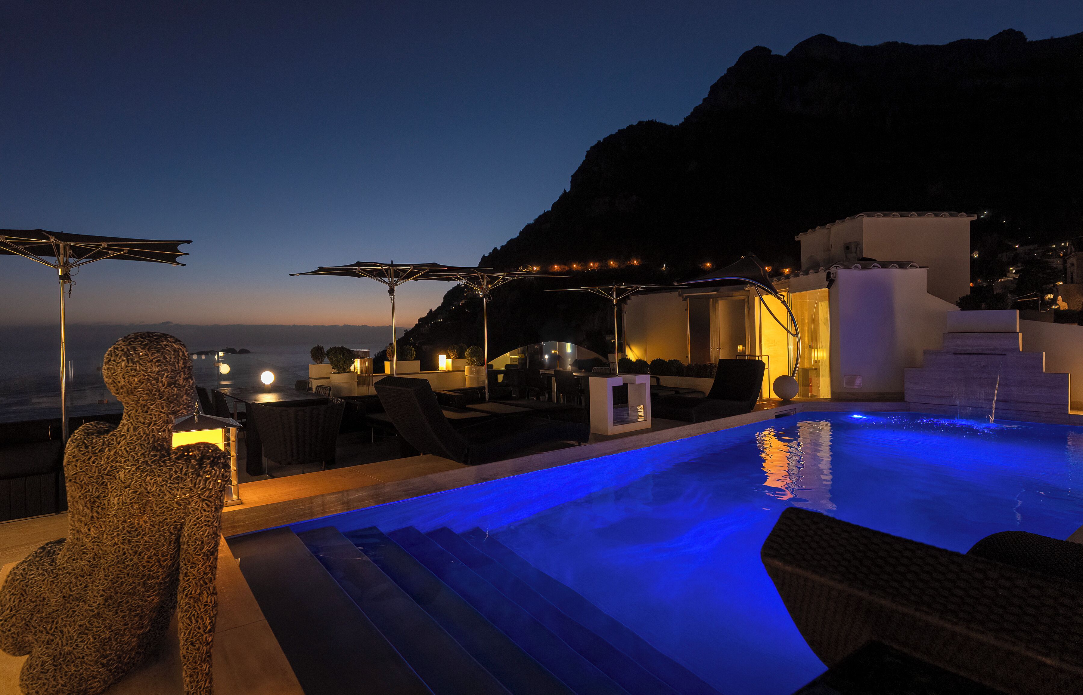 A poolside area with lounge chairs, a sculpture, and a view of the sea at dusk.