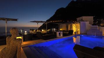 A poolside area with lounge chairs, a sculpture, and a view of the sea at dusk.