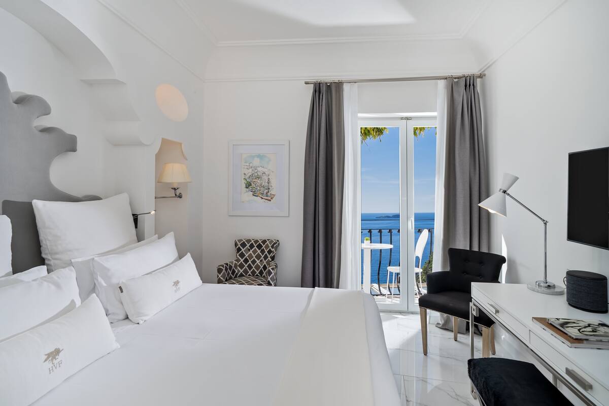 A hotel room with a bed, a desk, a chair, a TV, and a balcony with a view of the sea.