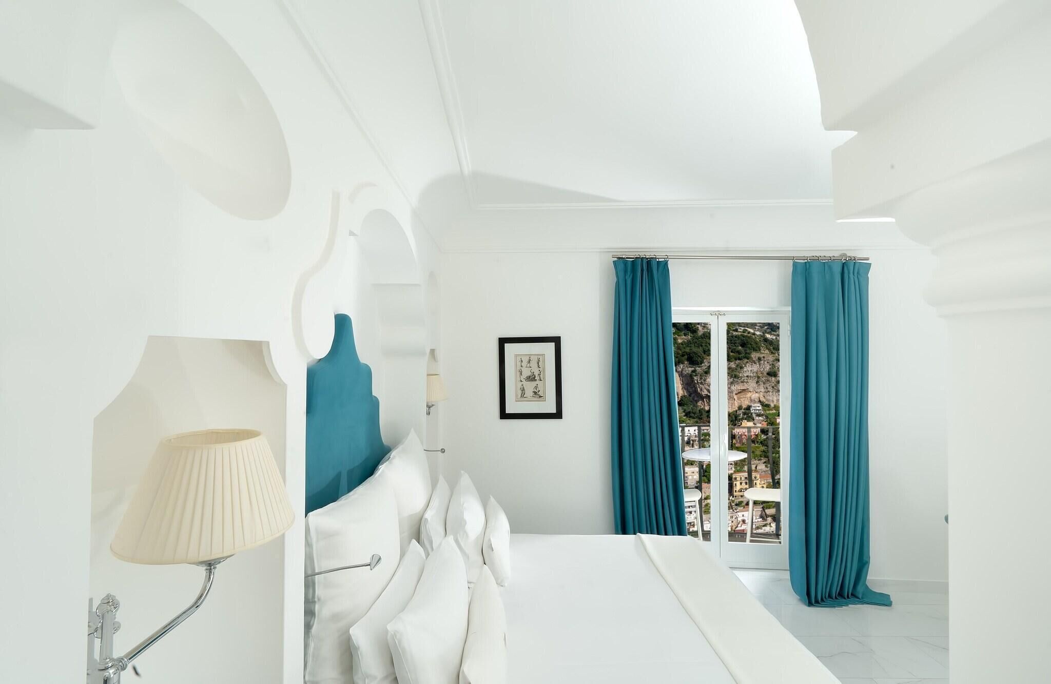 A bedroom with a bed, a lamp, a framed picture, and a view of a town through a window.