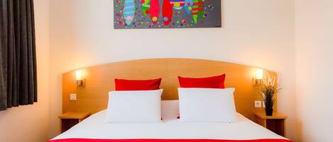 A hotel room with a large bed, red and white bedding, two red pillows, a colorful abstract painting on the wall, and bedside lamps.