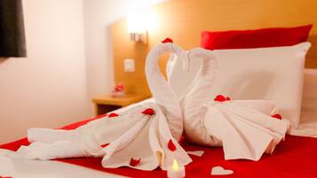 Una cama con cubrecama rojo y blanco, una toalla dispuesta en forma de corazón y una vela encendida.