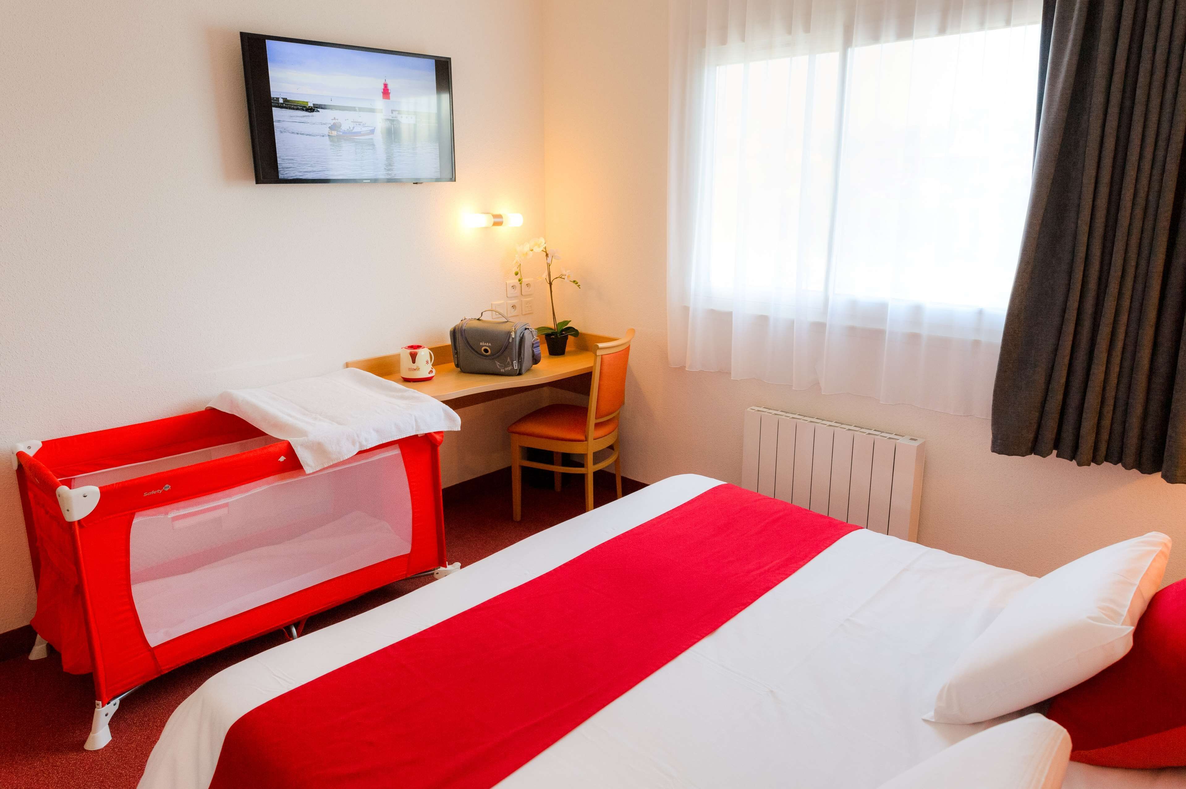 A hotel room with a bed, a desk, a chair, a red crib, a television on the wall, and a window with curtains.