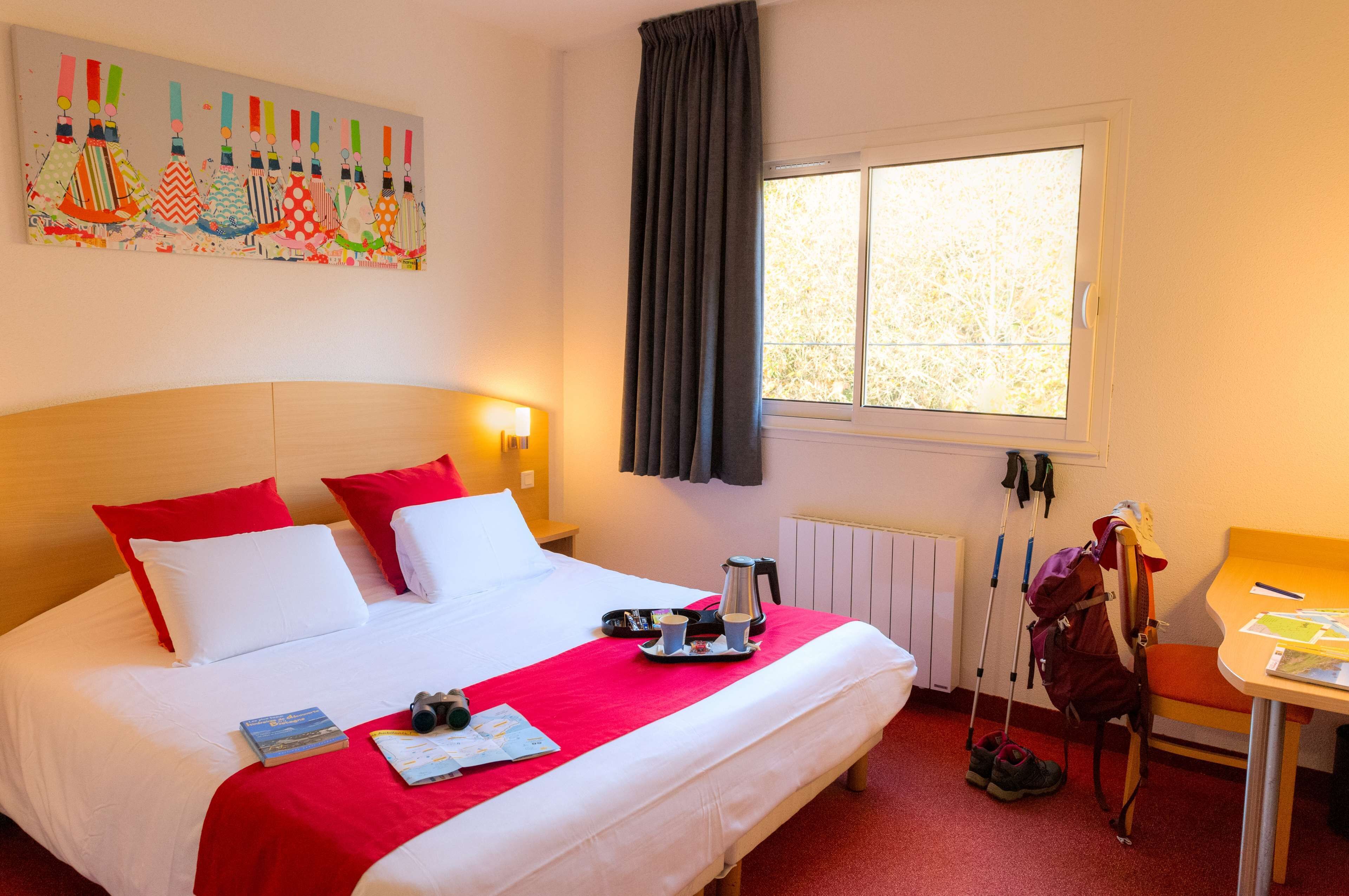 Habitación de hotel con cama, ropa de cama roja y blanca, una ventana con cortinas, una mesa con una silla y una mochila en el suelo.