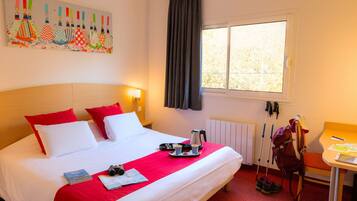 Habitación de hotel con cama, ropa de cama roja y blanca, una ventana con cortinas, una mesa con una silla y una mochila en el suelo.