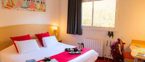Habitación de hotel con cama, ropa de cama roja y blanca, una ventana con cortinas, una mesa con una silla y una mochila en el suelo.