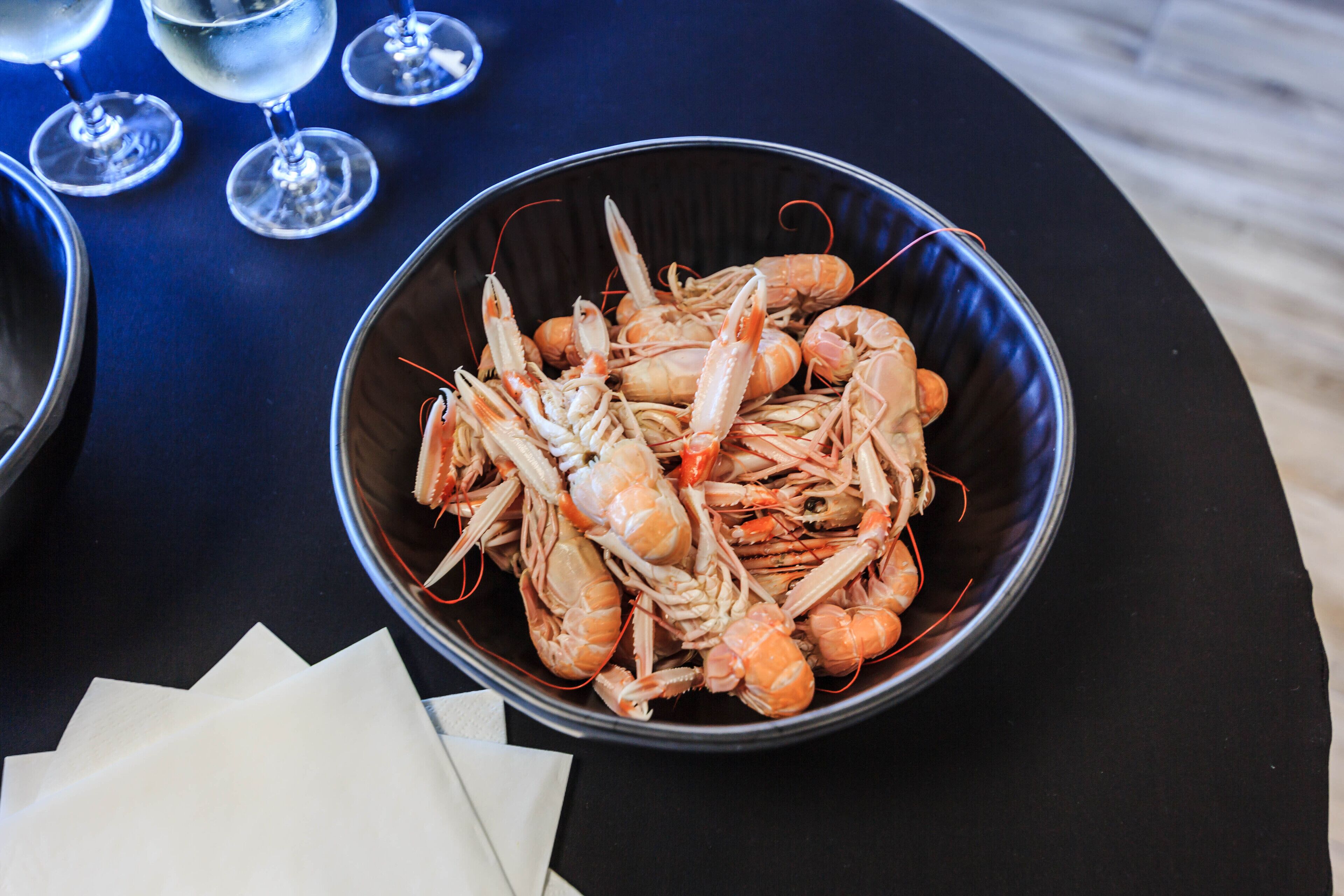A bowl of cooked shrimp on a table with a napkin and wine glasses in the background.