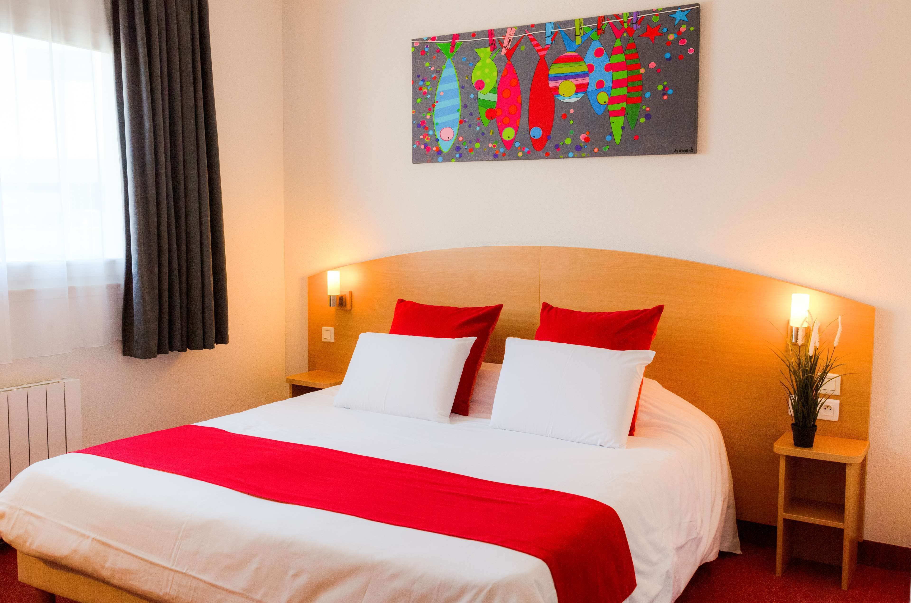 A hotel room with a large bed, red and white bedding, a wooden headboard, bedside lamps, and a wall-mounted artwork.