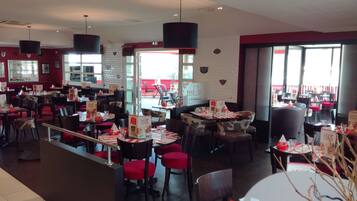 A restaurant interior with multiple tables and chairs, red cushioned chairs, and black pendant lights.