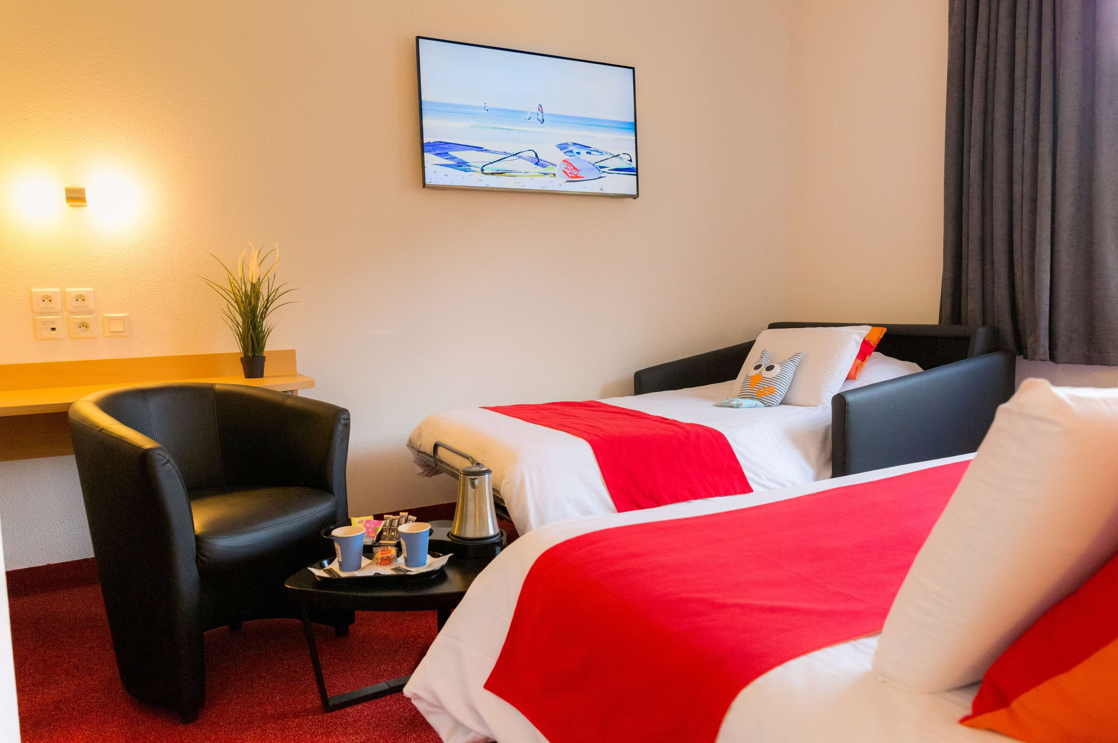 Une chambre d’hôtel avec deux lits, une chaise, une petite table avec un plateau, un tableau représentant une scène de plage et une plante en pot.