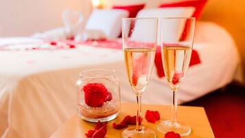 Un lit avec une literie blanche et des oreillers rouges, deux flûtes à champagne et un bocal en verre contenant une rose rouge.