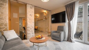A living room with a stone wall, a grey sofa, a round wooden coffee table with fruit, a grey armchair, and a TV mounted on the wall. - Villa Matejuska Heritage (Split)