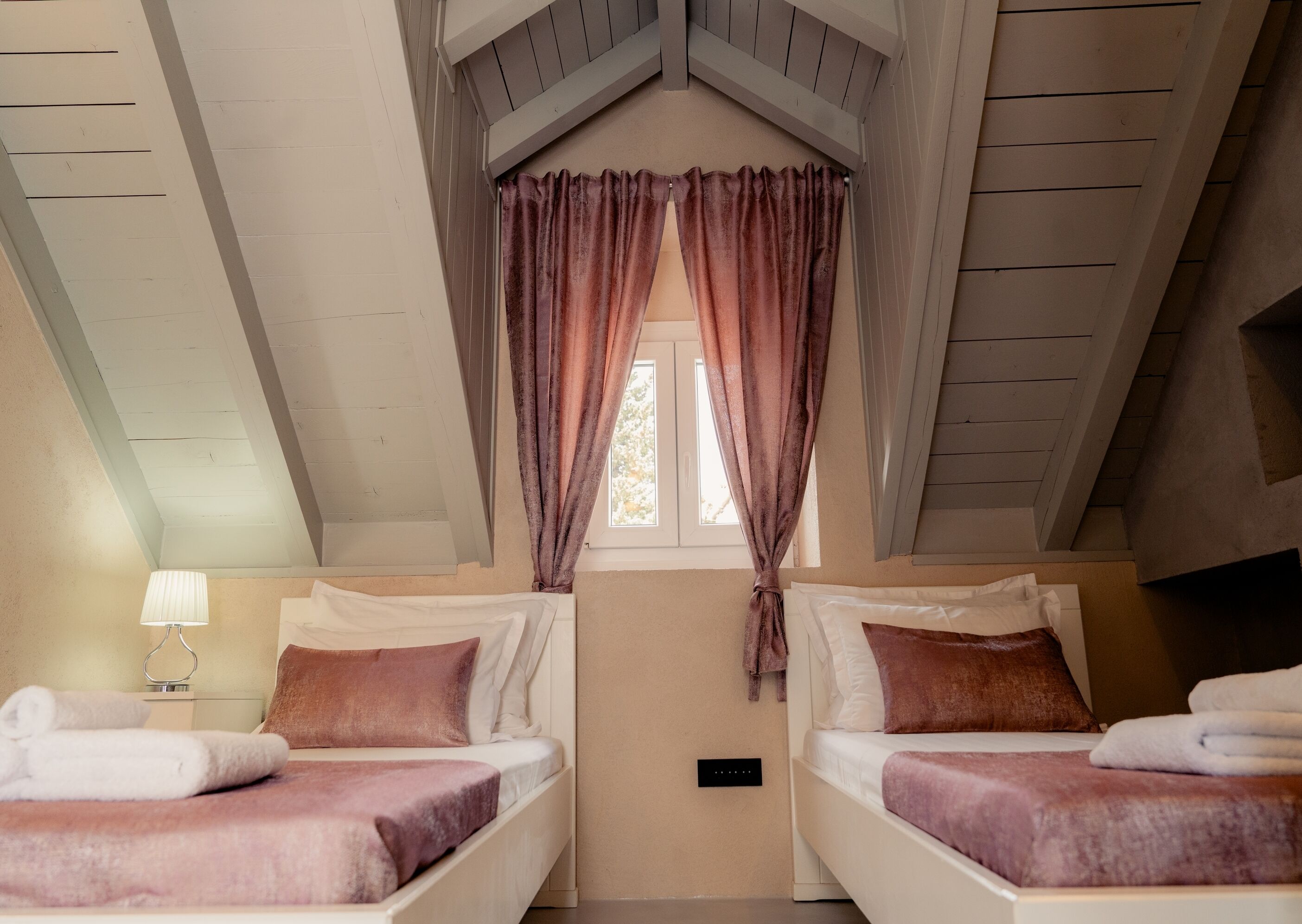 Una habitación con dos camas, una ventana con cortinas, una lámpara y un interruptor de pared.