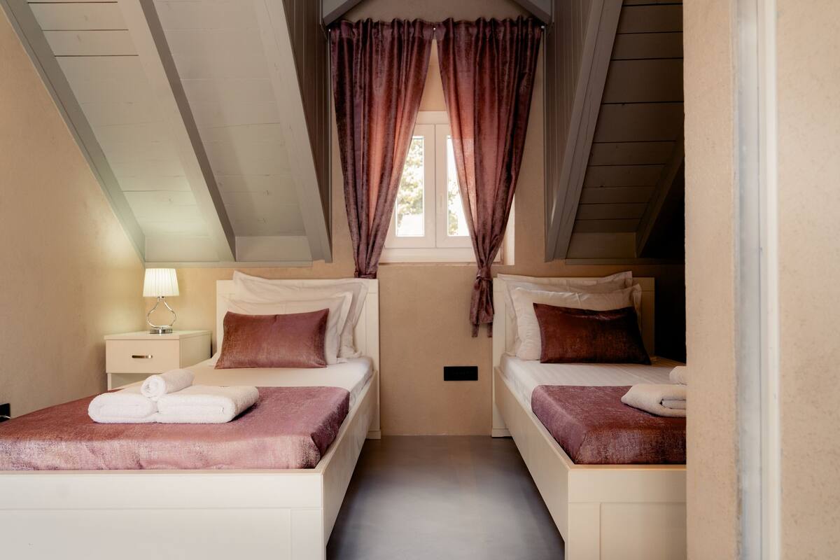 A room with two beds, a nightstand with a lamp, and a window with curtains.