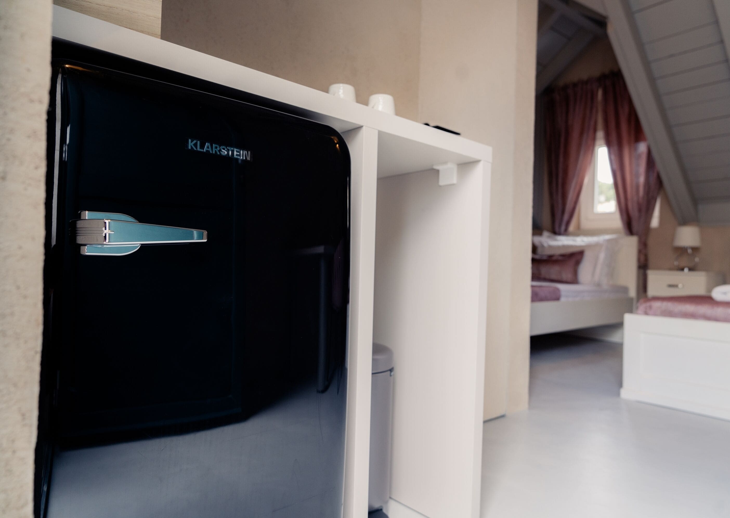 Une chambre moderne avec un mini-bar noir, des murs blancs et un lit doté d’une tête de lit.