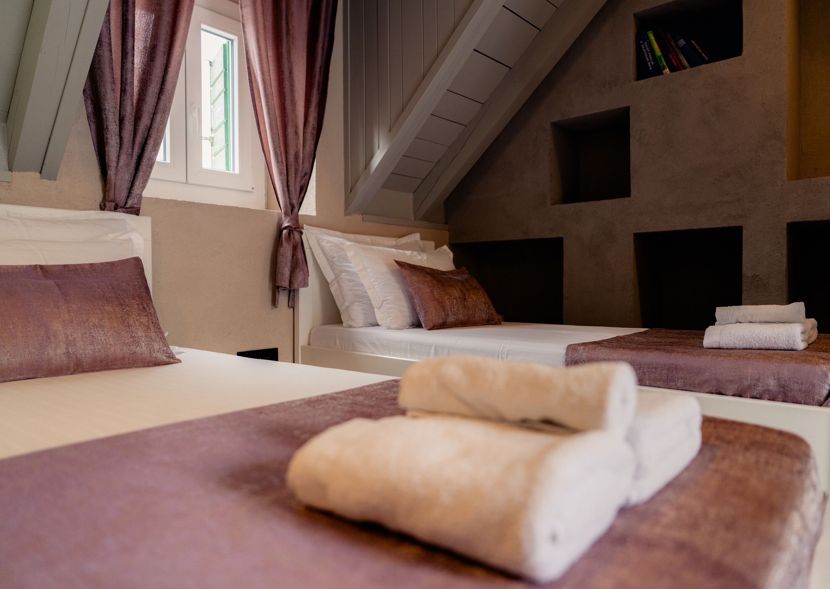 Een hotelkamer met twee bedden, een raam met gordijnen, een boekenkast en een schuine plafond.