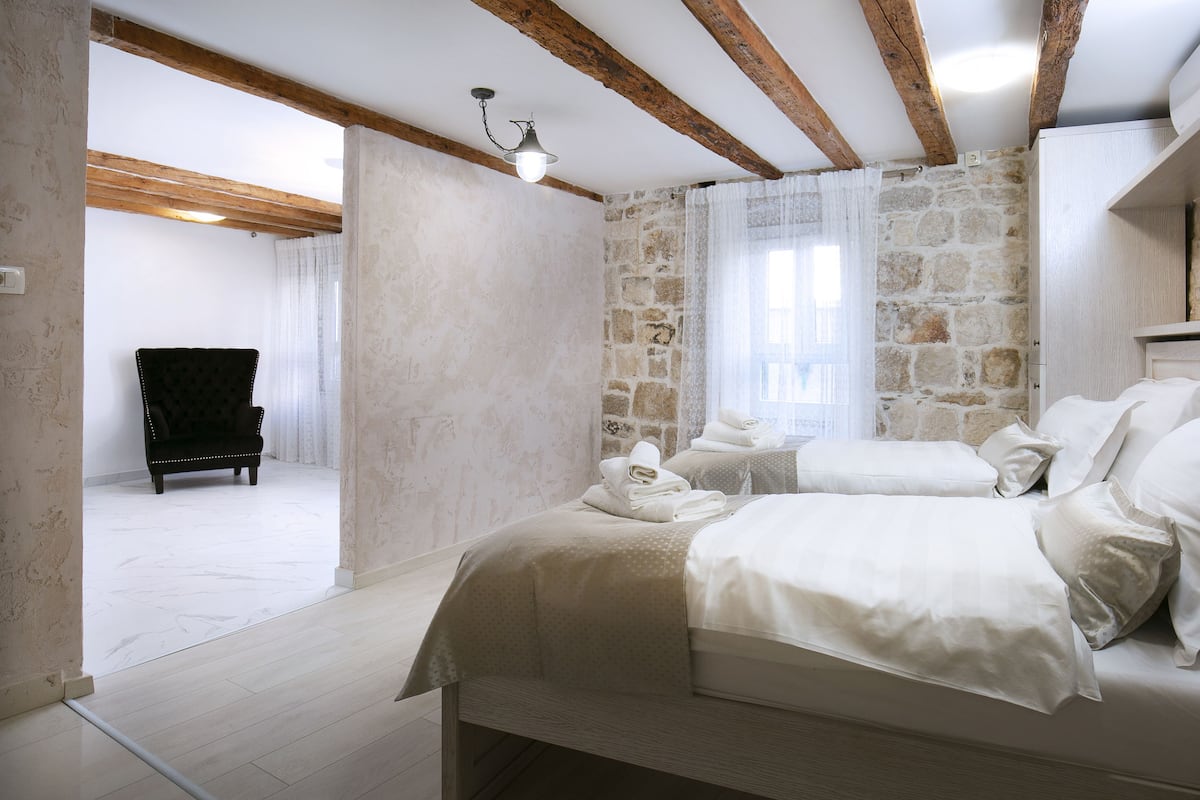 A bedroom with a stone wall, a bed with white linens, a wooden chair, and a small table with towels.