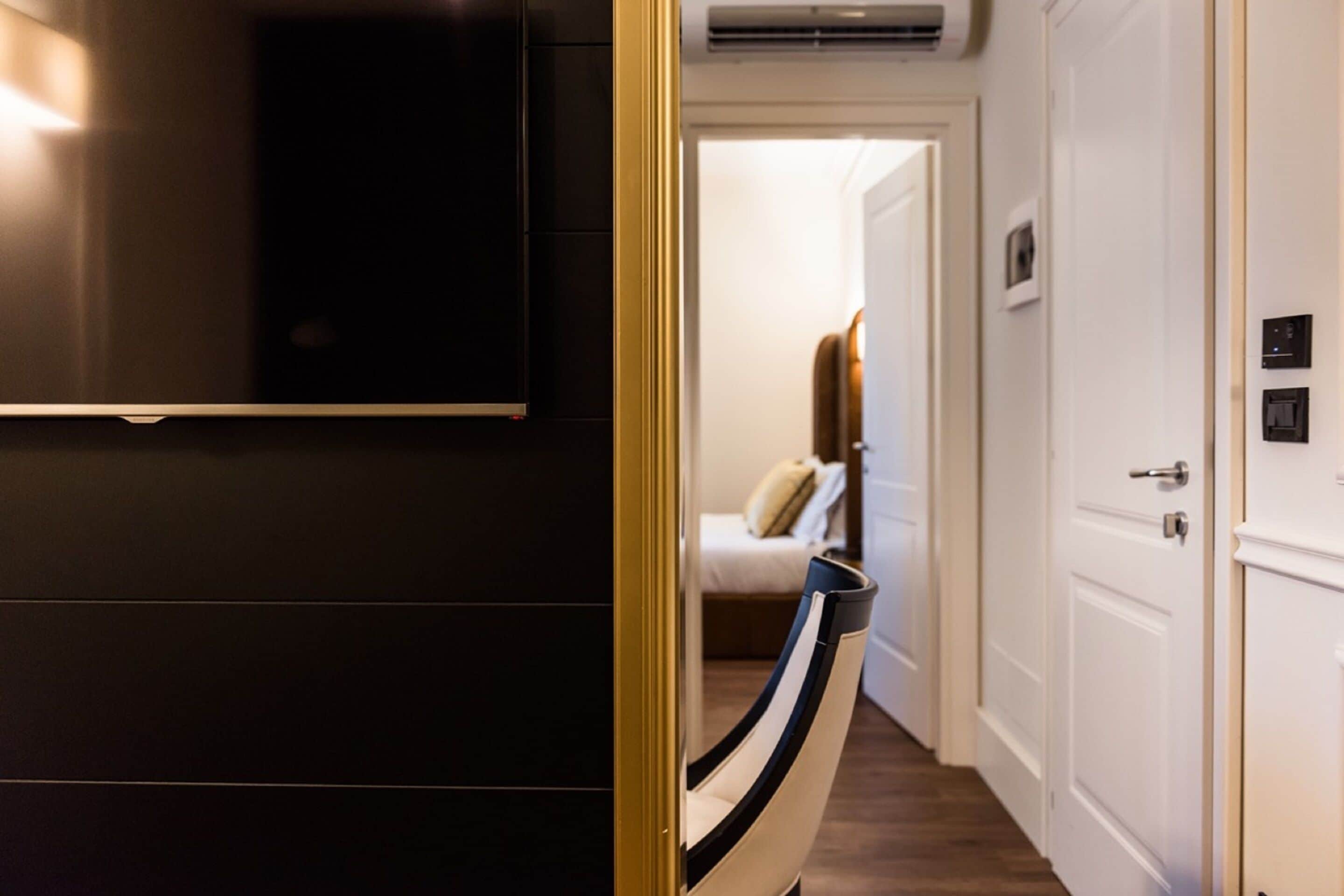 Quarto de hotel com um guarda-roupa escuro, uma televisão na parede, uma cama com cabeceira branca e uma porta que dá para outro quarto.