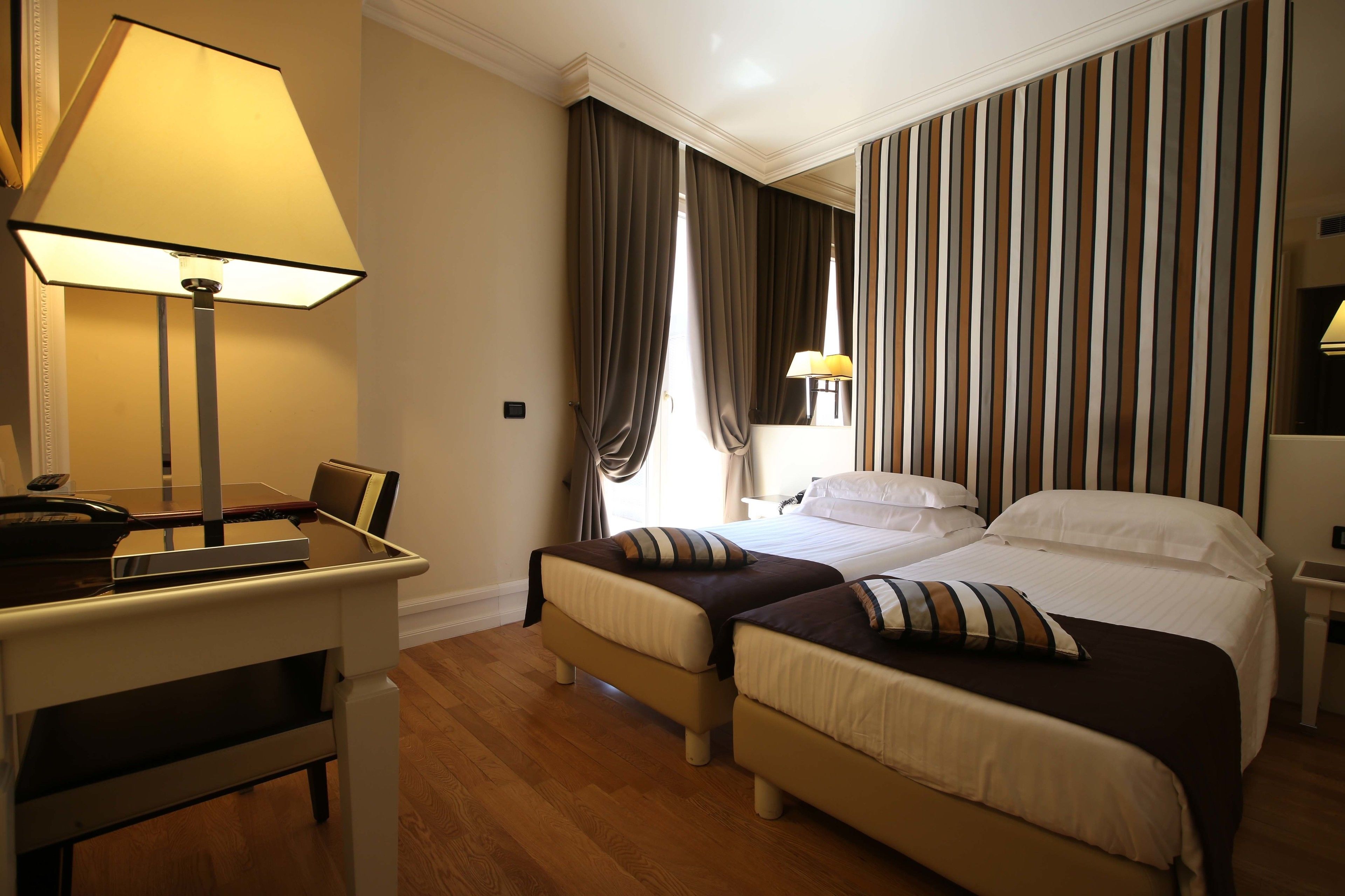 Una habitación de hotel con dos camas, un escritorio con lámpara y un ventanal con cortinas.