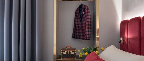 A hotel room with a bed, red pillows, a mirror with a coat, and a suitcase on a nightstand.