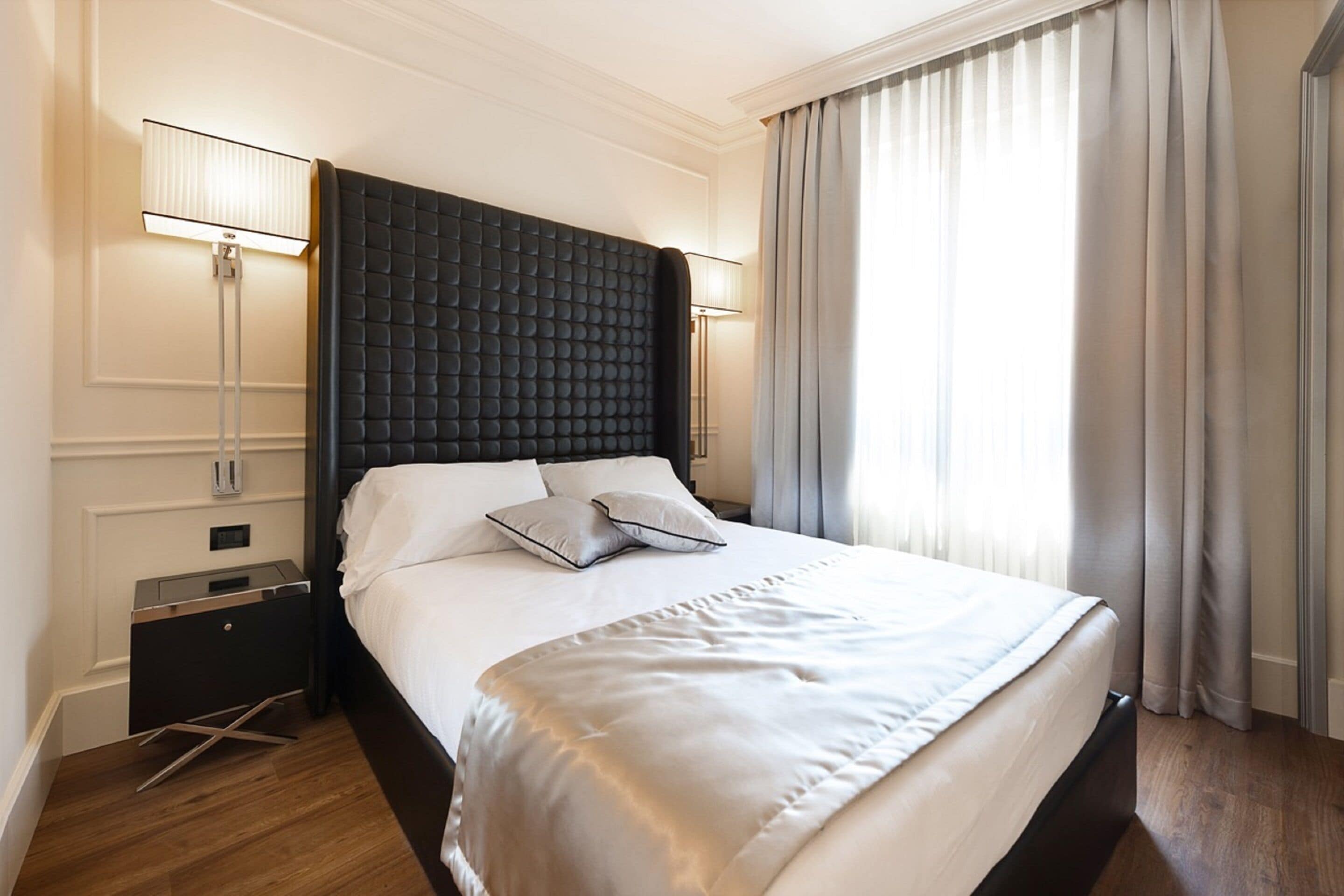 Habitación de hotel moderna con una cama grande, cabecera oscura, lámparas de noche y vistas al exterior a través de cortinas.