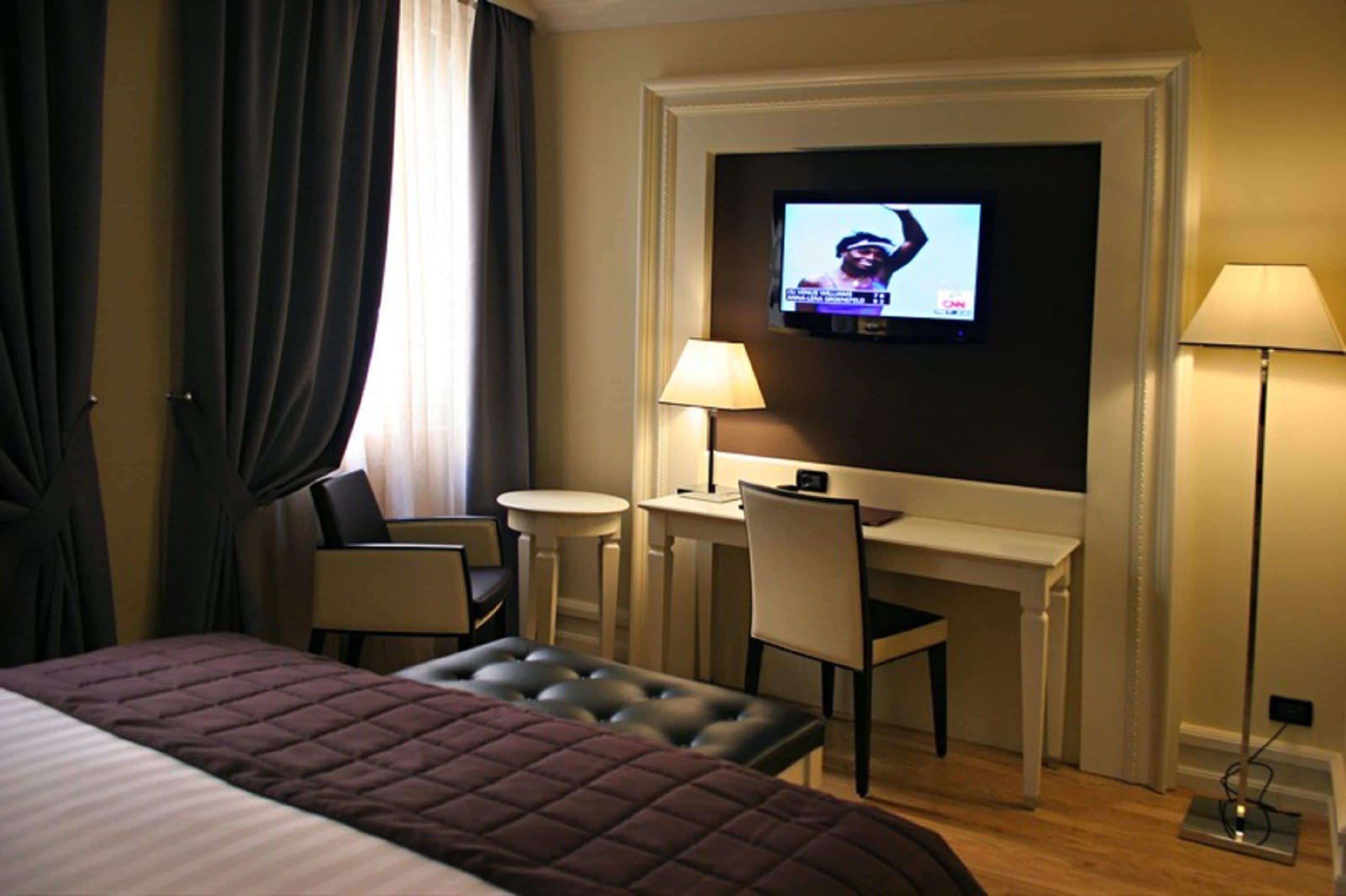 A hotel room with a bed, a TV mounted on the wall, a desk with a chair, a small table, and a lamp.