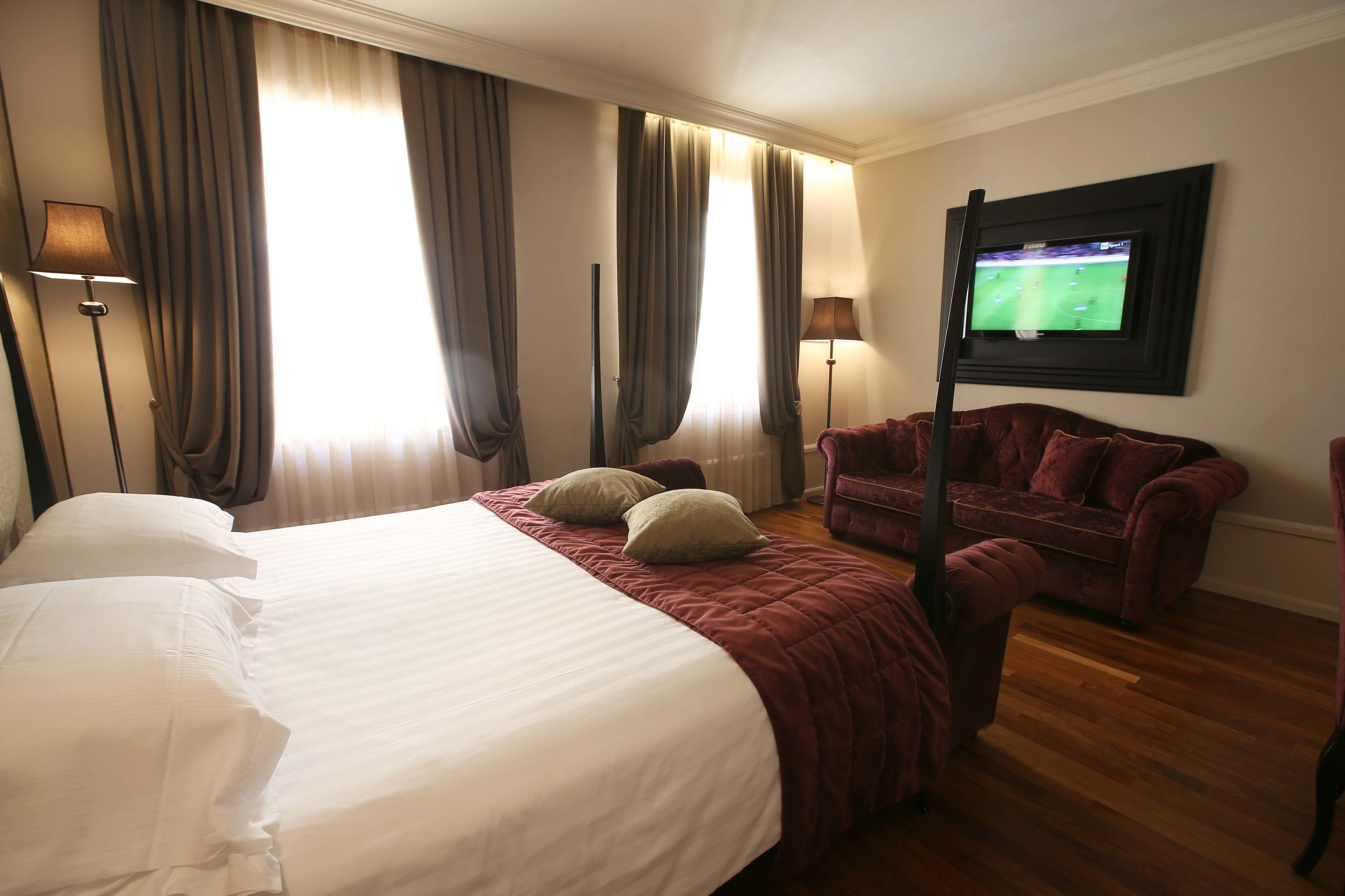 Habitación de hotel con una cama grande, dos almohadas, un sillón rojo, un televisor en la pared y cortinas en las ventanas.