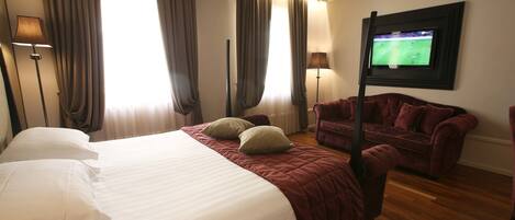 Habitación de hotel con una cama grande, dos almohadas, un sillón rojo, un televisor en la pared y cortinas en las ventanas.