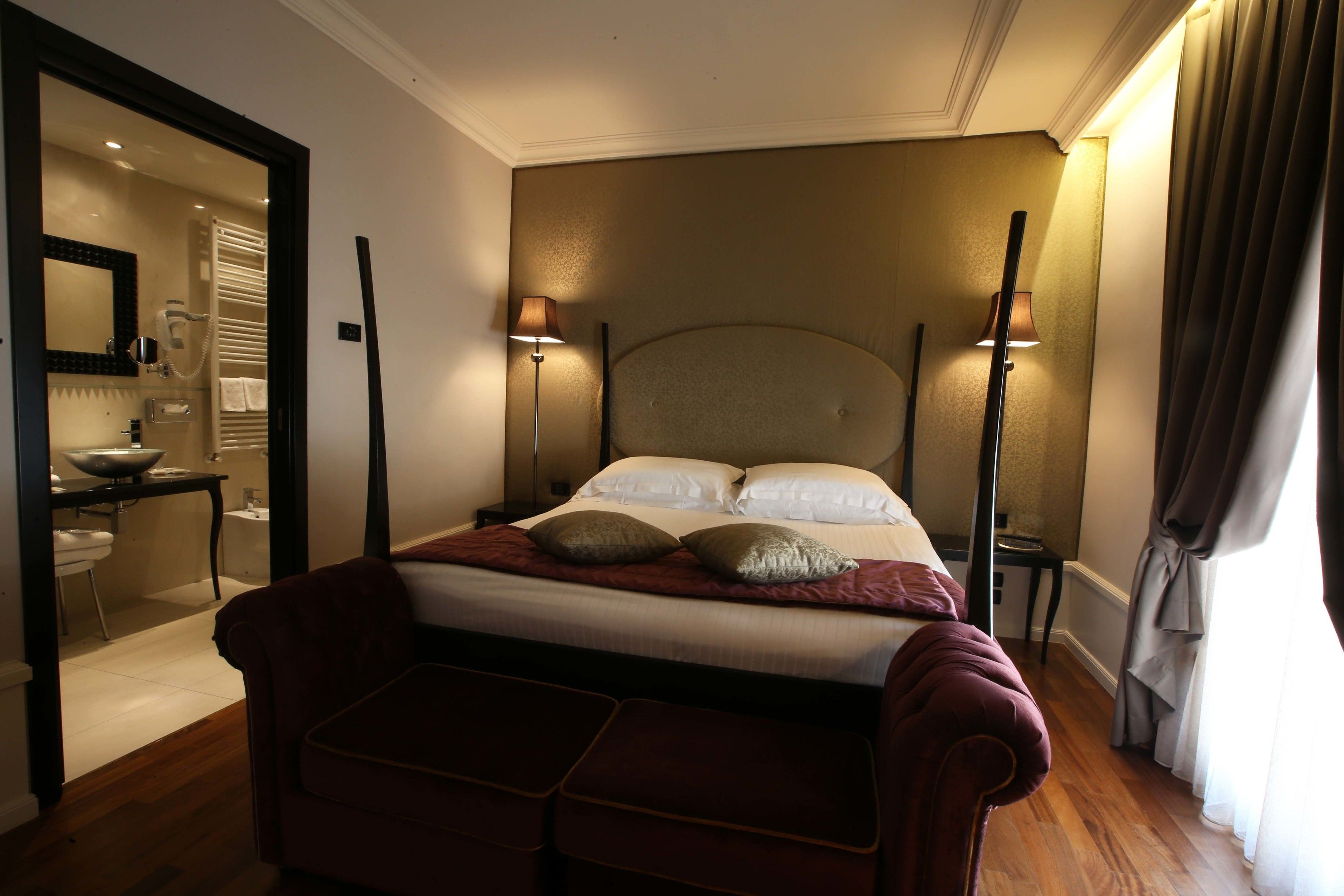 Ein Hotelzimmer mit Bett, Nachttischlampen, einem Spiegel und einem durchgegangenen Tür angedeuteten Badezimmer.