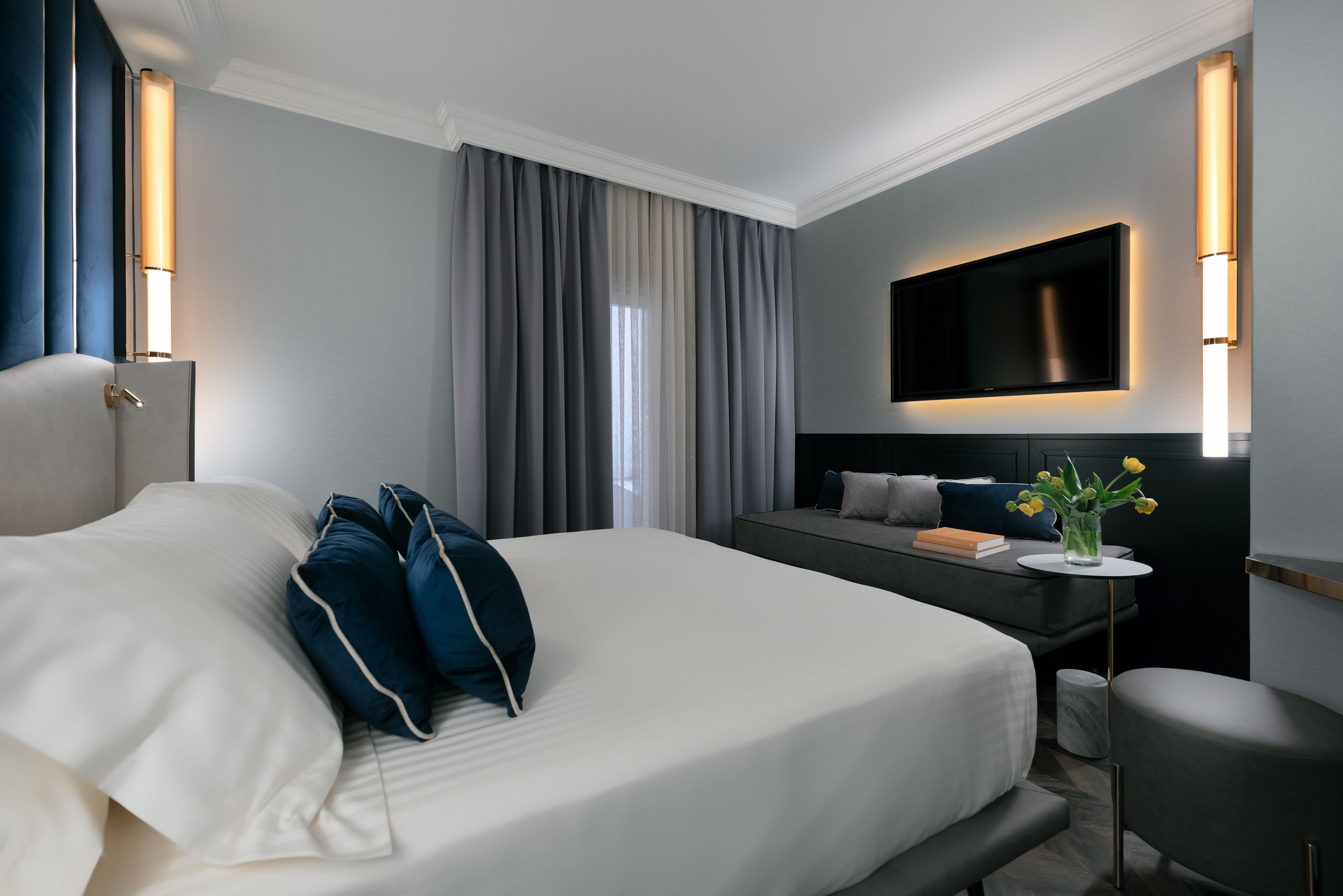 A modern hotel room with a large bed, a sofa, a TV mounted on the wall, and a small table with a vase of flowers.