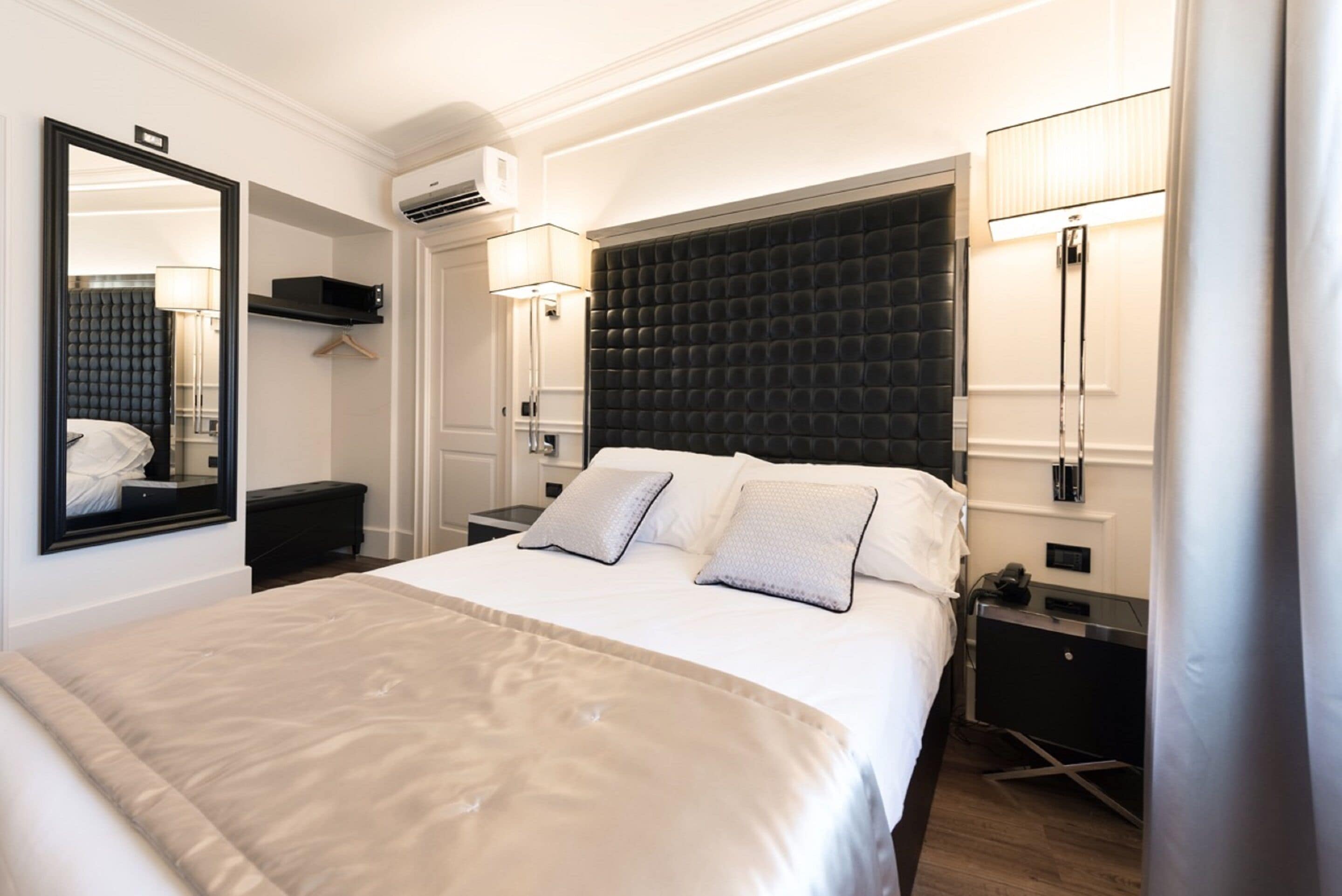 Una habitación de hotel moderna con una cama grande, un cabecero oscuro, lámparas de noche, un espejo, una mesita de noche y un equipo de aire acondicionado.