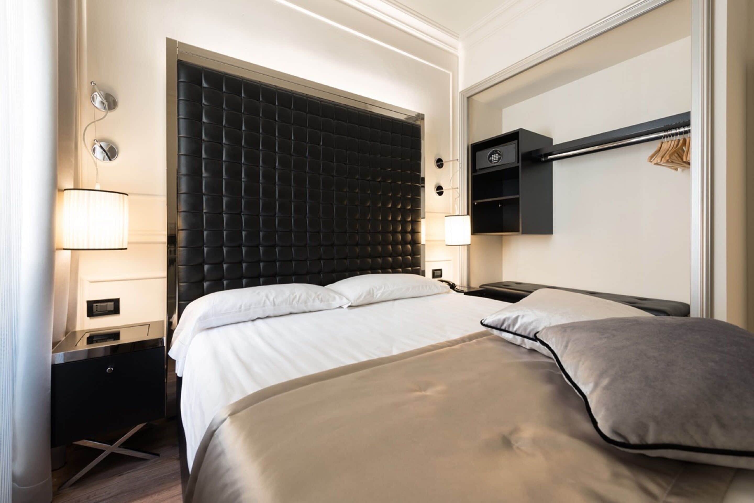 A hotel room with a large bed, a bedside table with a lamp, a wardrobe, and a wall-mounted shelf.