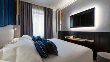 Habitación de hotel con una cama grande, una mesita de noche con lámpara, un televisor montado en la pared y una ventana con cortinas.