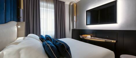A hotel room with a large bed, a bedside table with a lamp, a TV mounted on the wall, and a window with curtains.