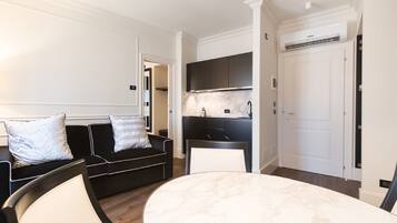 A modern living room with a black sofa, a round marble table, and a kitchen area with dark cabinets and a white door.