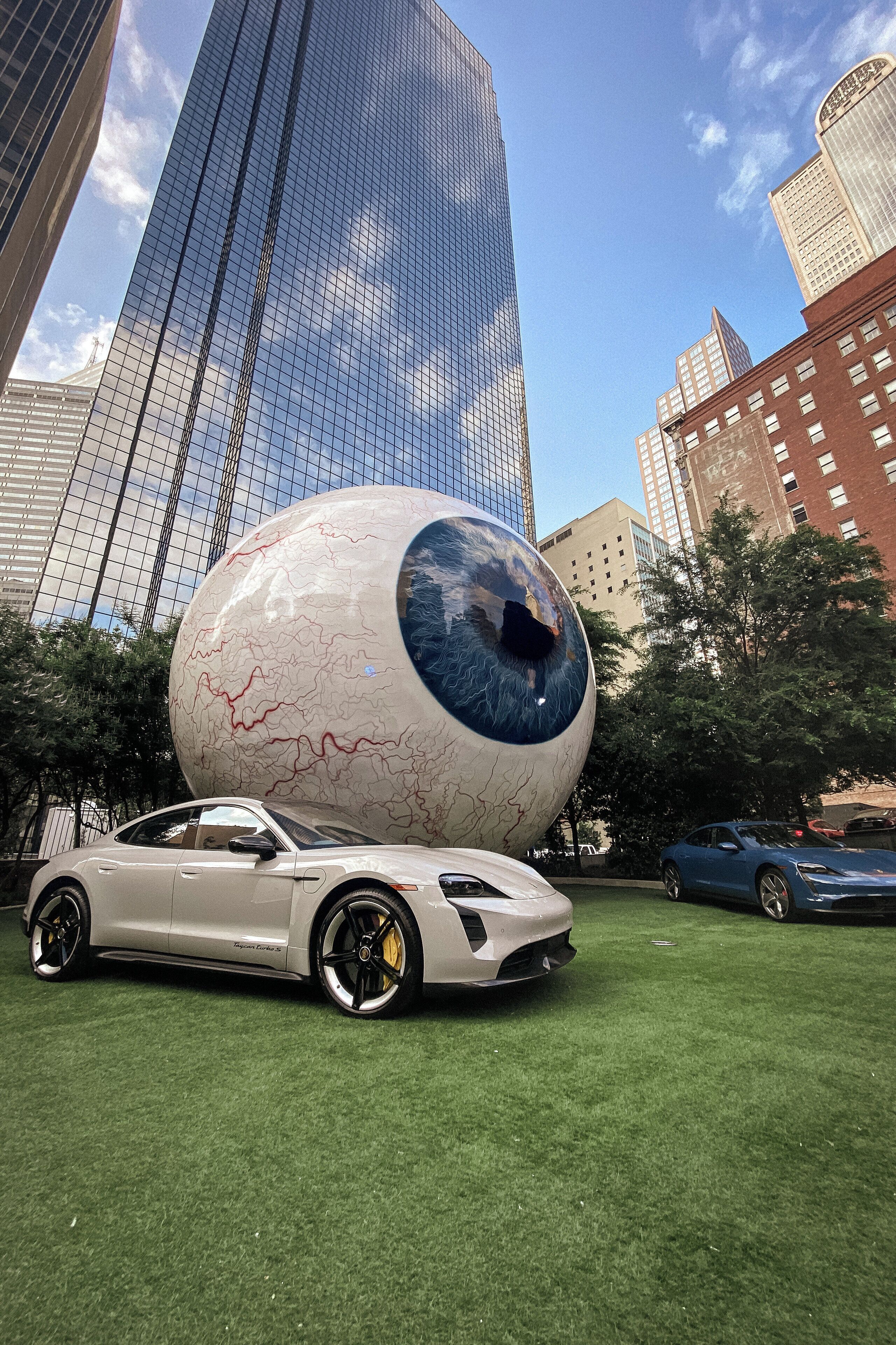 A modern building with a glass facade, a large spherical sculpture, and a sports car parked on a grassy area.