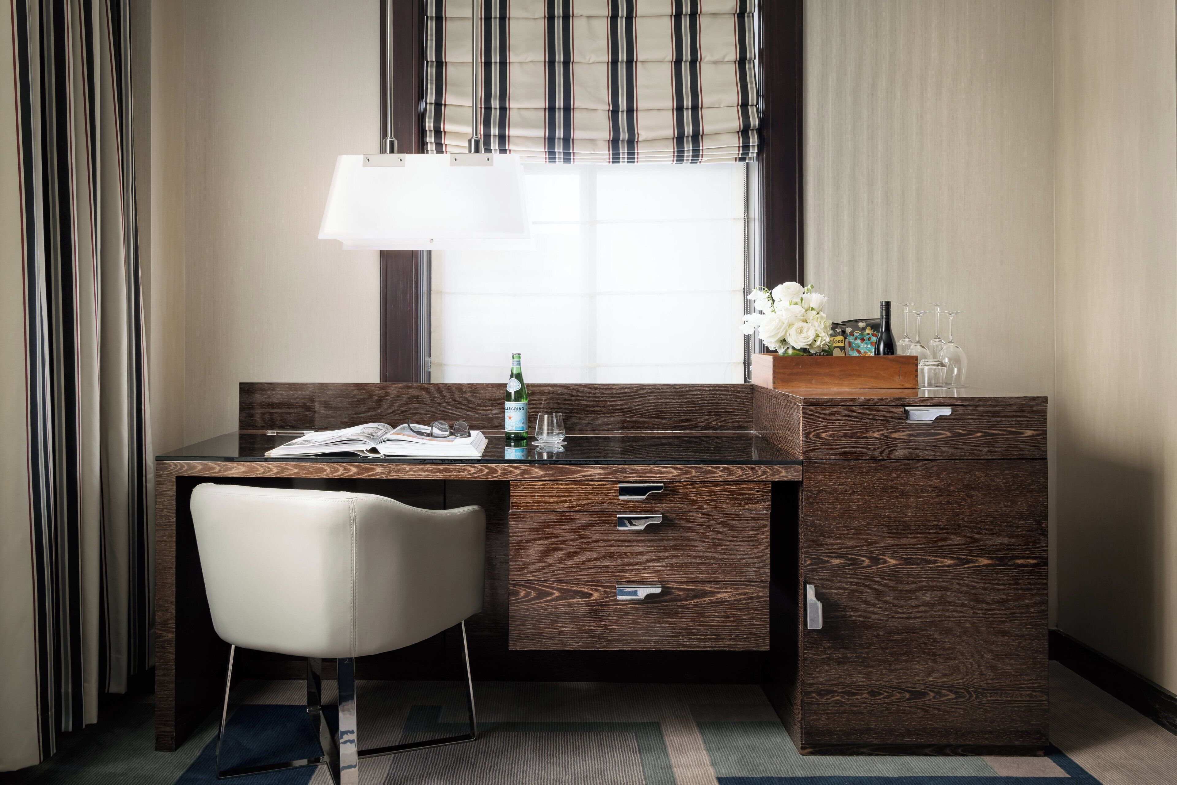 A hotel room with a wooden desk, a white chair, a lamp, a bottle of water, a glass, and a vase with flowers.