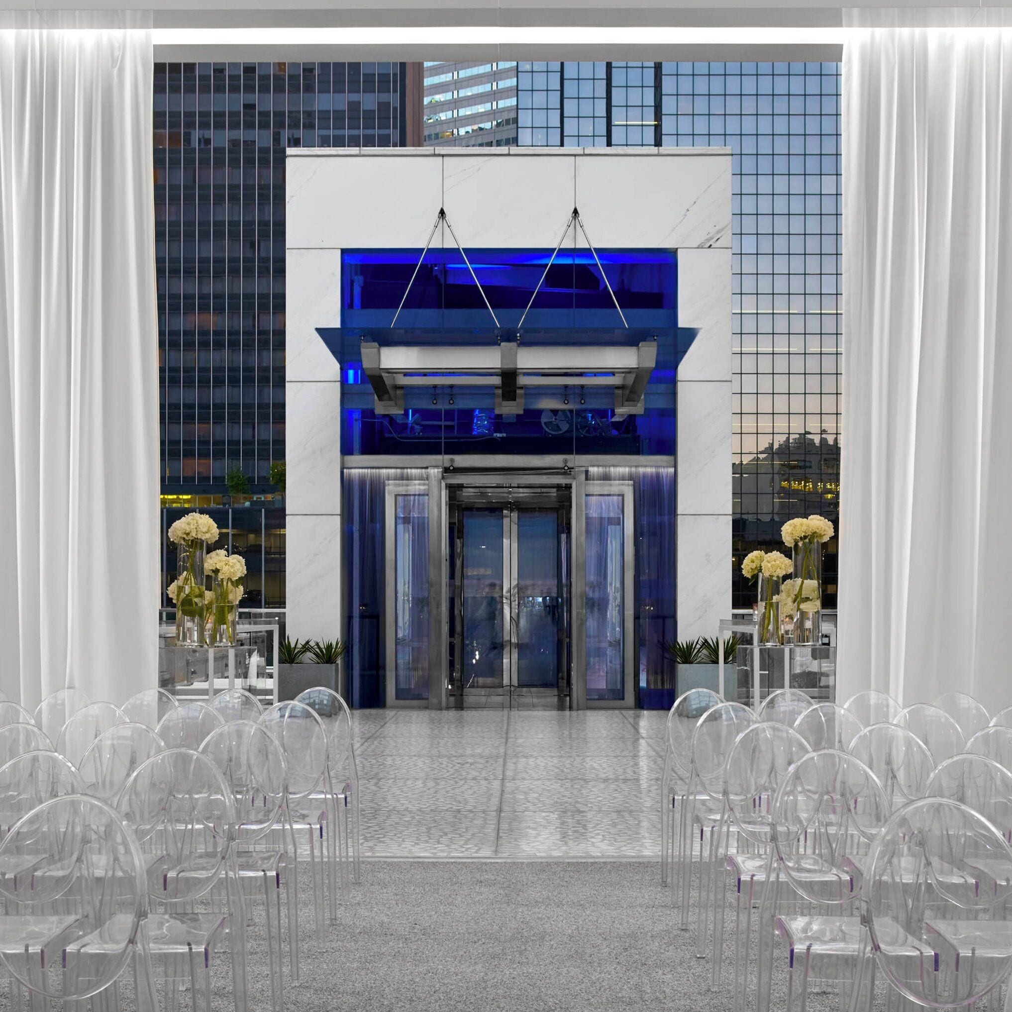 A modern hotel entrance with a glass facade, white curtains, and clear chairs arranged for an event.