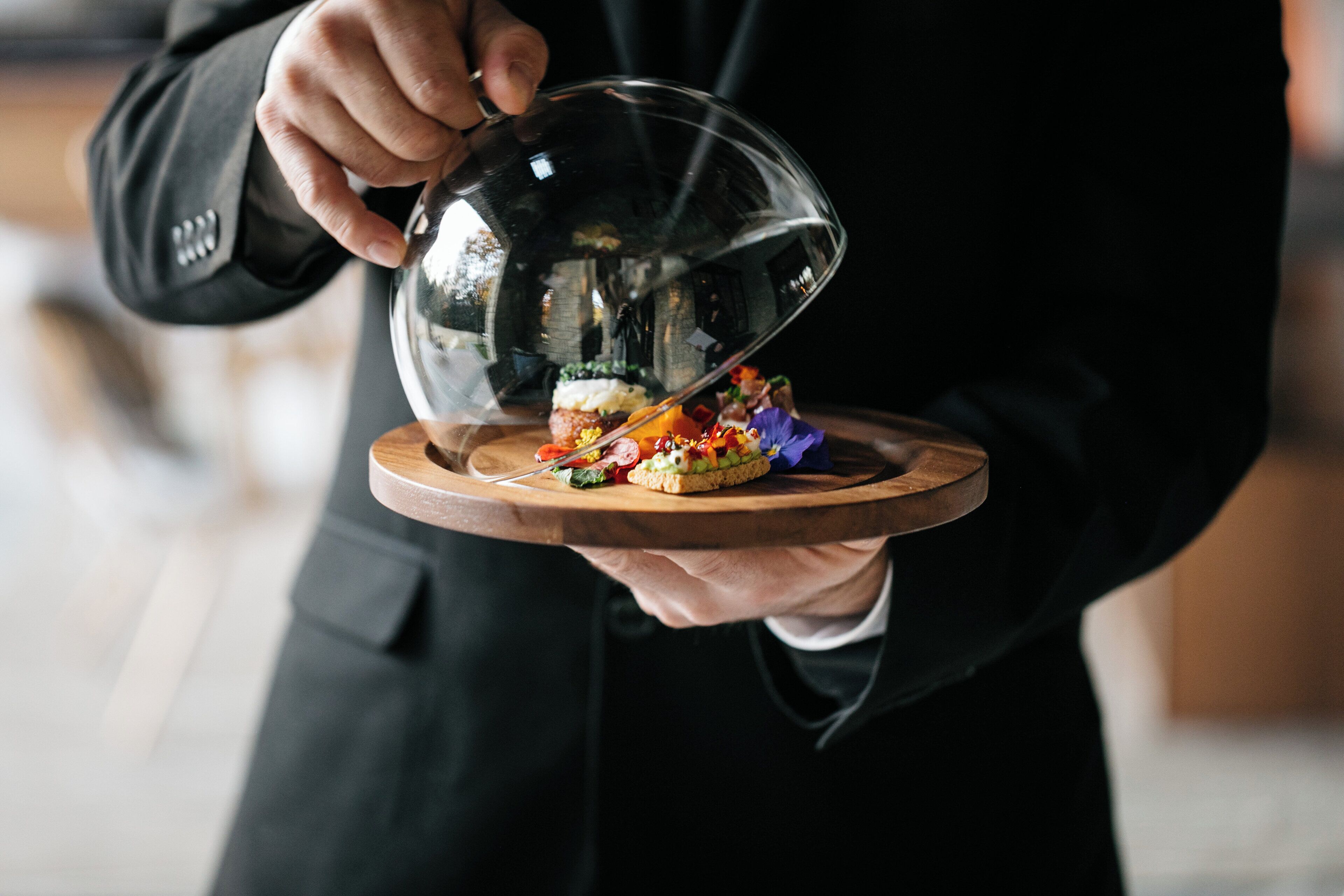 A person holding a wooden board with a food item under a dome-like cover.