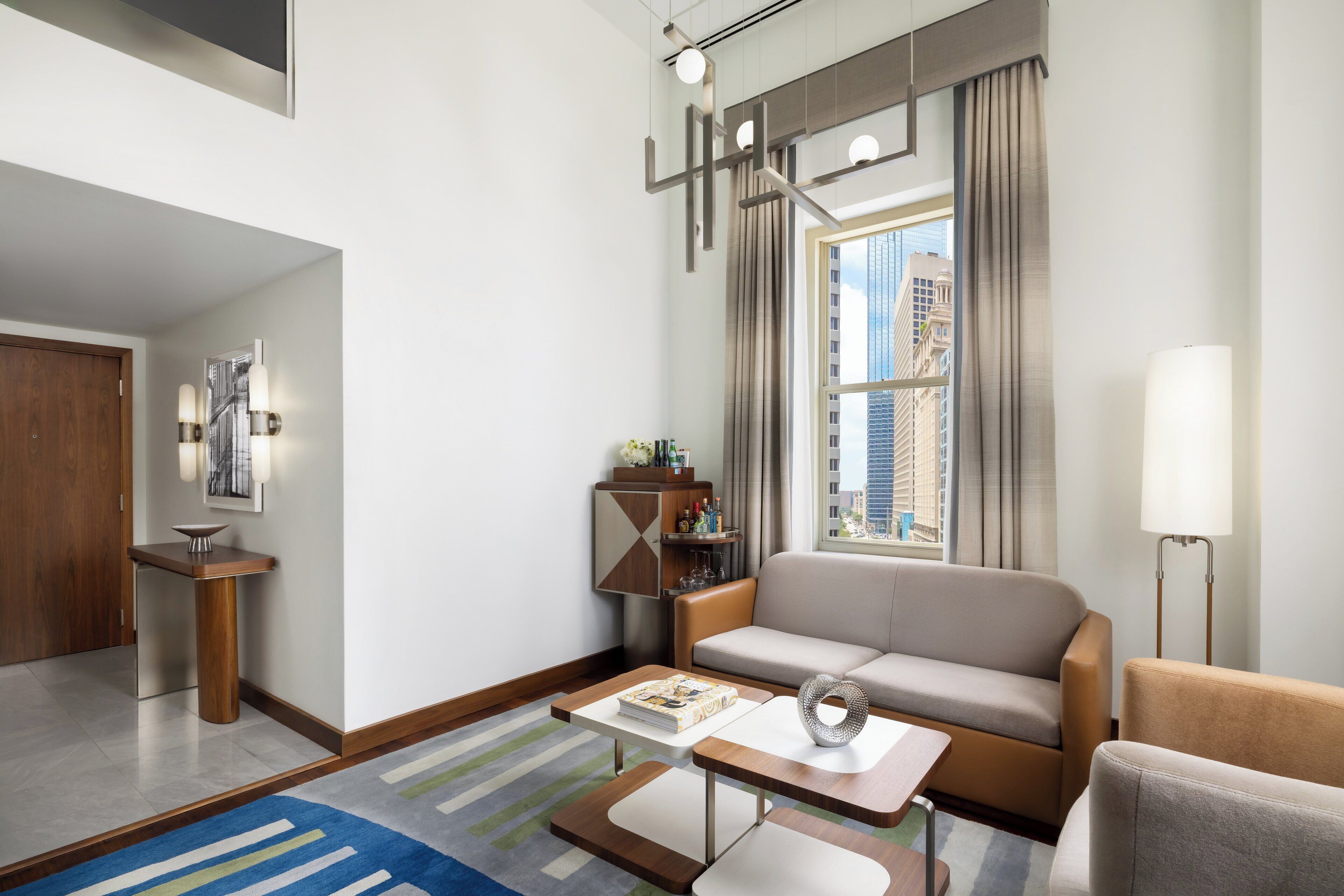A modern living room with a sofa, coffee table, and a lamp. There is a window with curtains and a view of buildings.