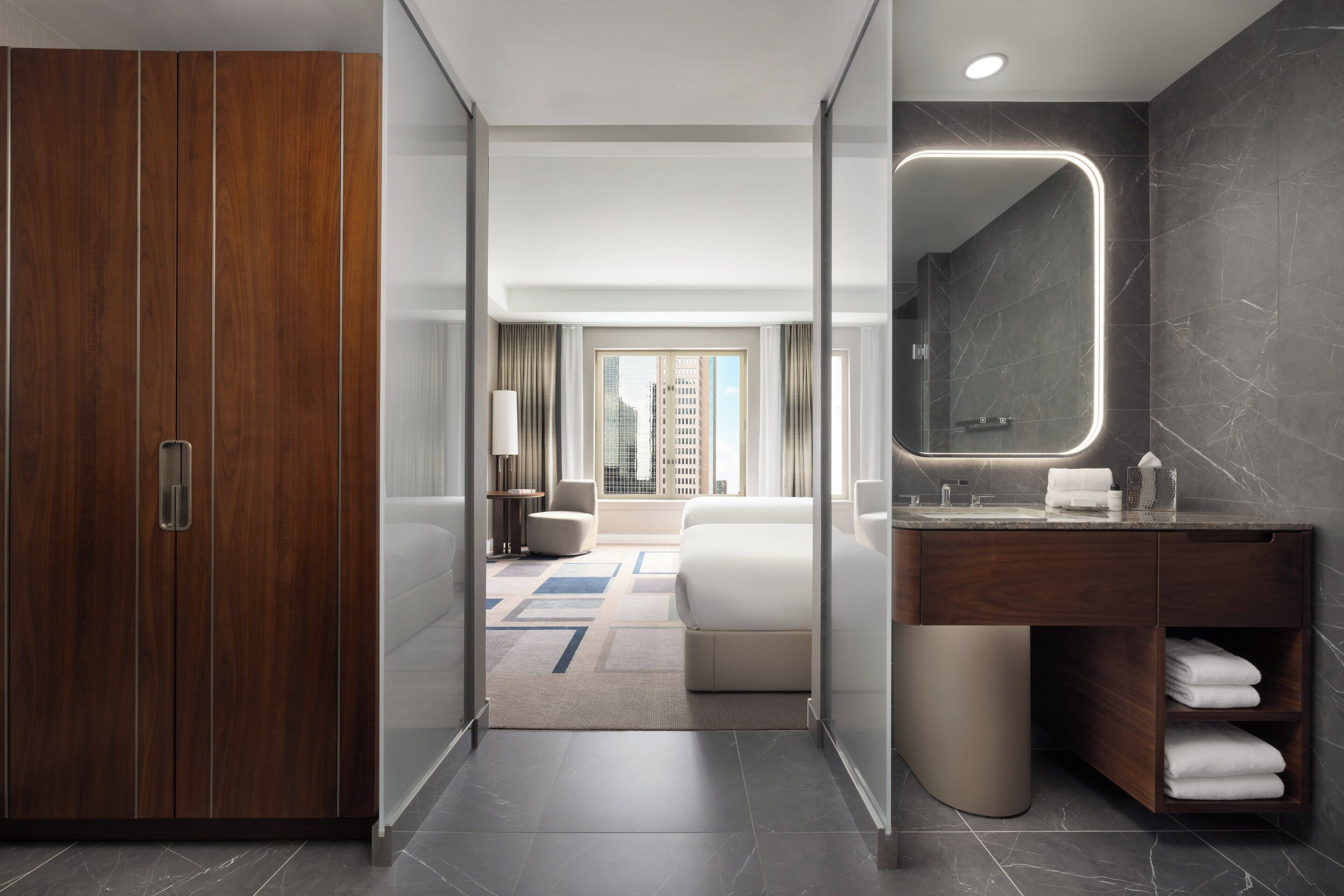 A modern hotel room with a wooden wardrobe, a large mirror, a bathroom with a marble countertop, and a view of a cityscape.