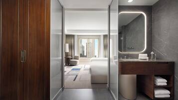 A modern hotel room with a wooden wardrobe, a large mirror, a bathroom with a marble countertop, and a view of a cityscape.