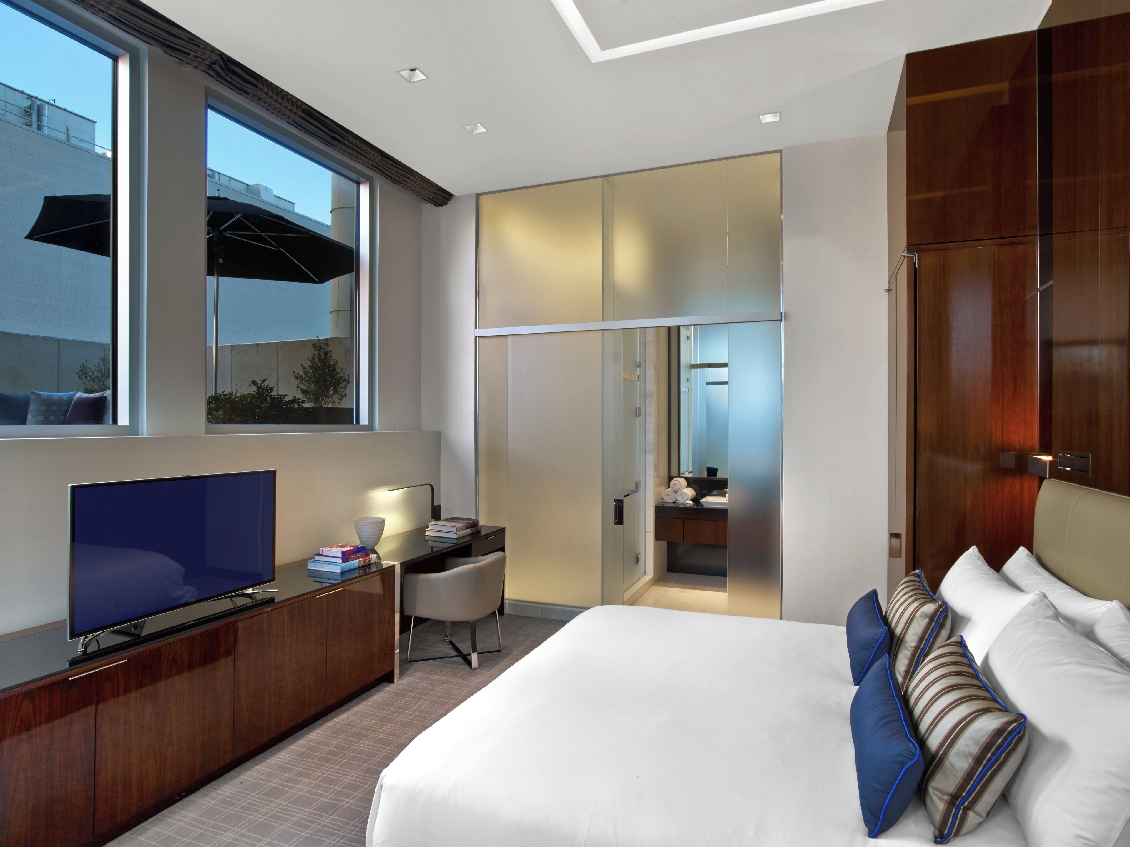 A modern hotel room with a large bed, a TV, a desk with a chair, and a bathroom visible through a glass door.