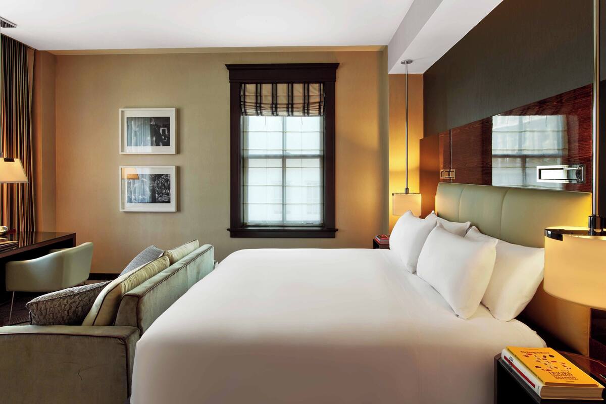 Habitación de hotel con una cama grande, un escritorio, una silla, un ventanal con persianas y dos cuadros enmarcados en la pared.