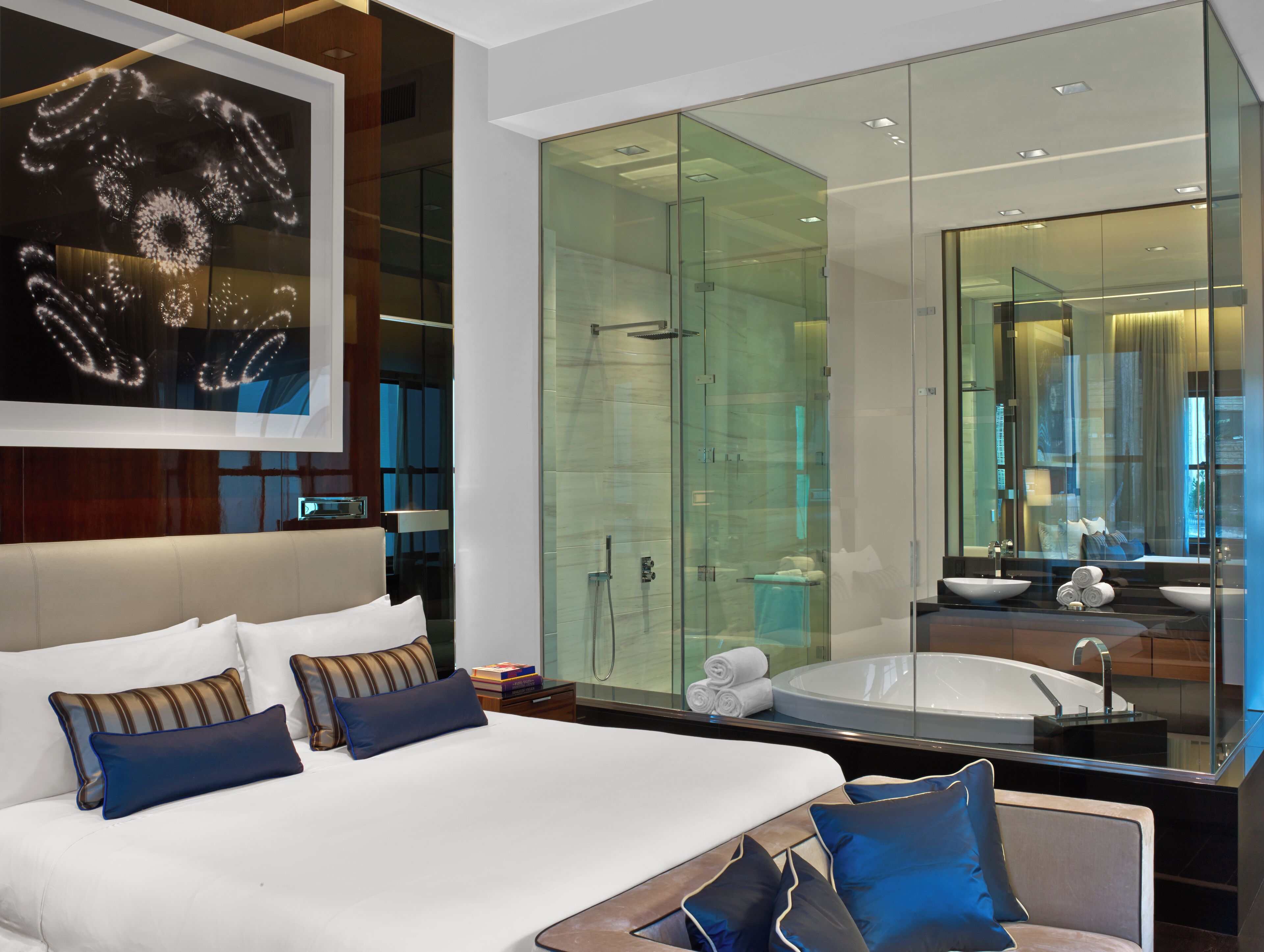 Ein modernes Hotelzimmer mit einem großen Bett, einer in Glas eingefassten Dusche und einem großen Spiegel, der das Badezimmer reflektiert.