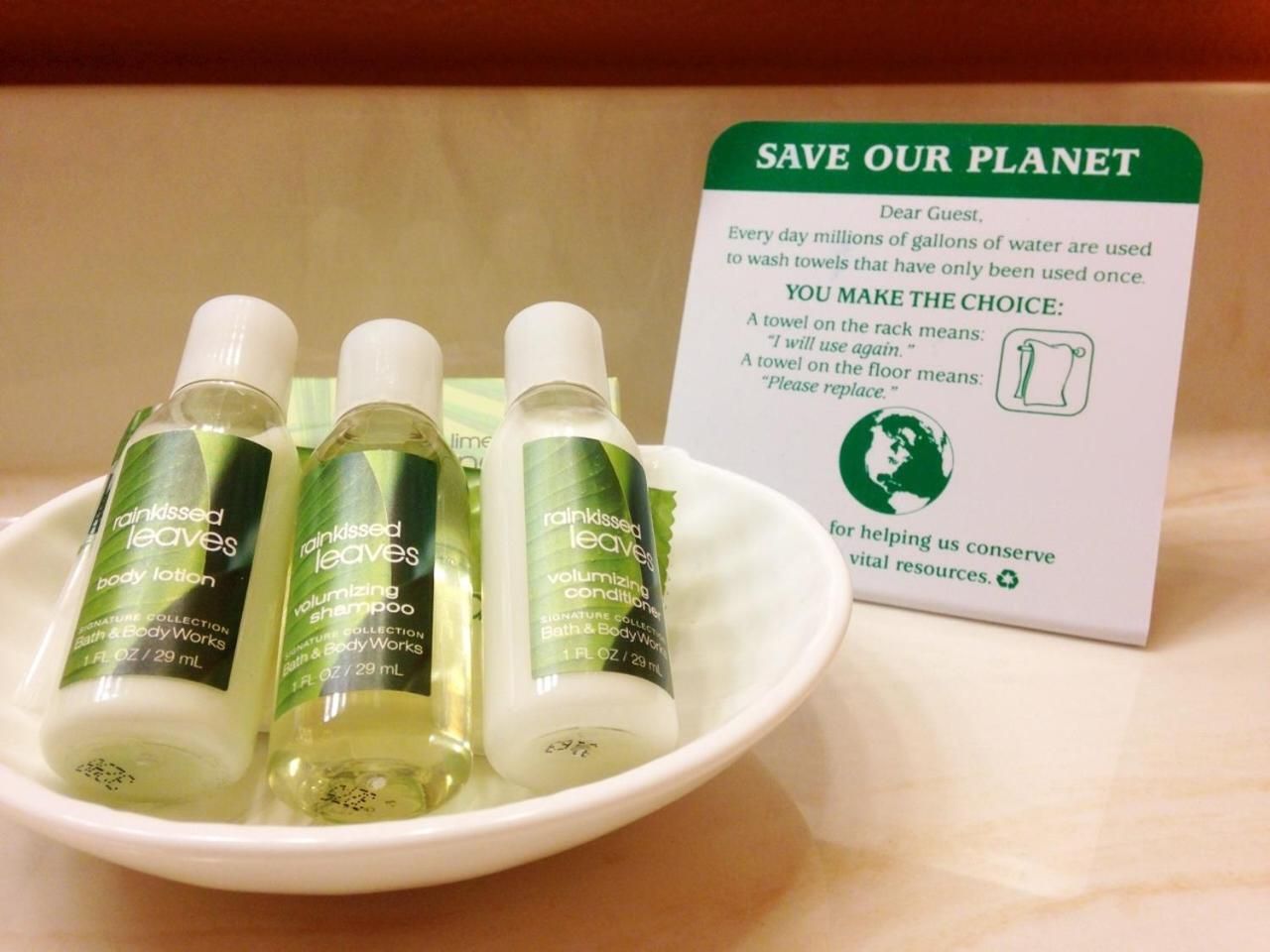 A white bowl containing three bottles of Rainkissed Leaves body lotion and shampoo, and a 'SAVE OUR PLANET' sign with instructions on towel usage.