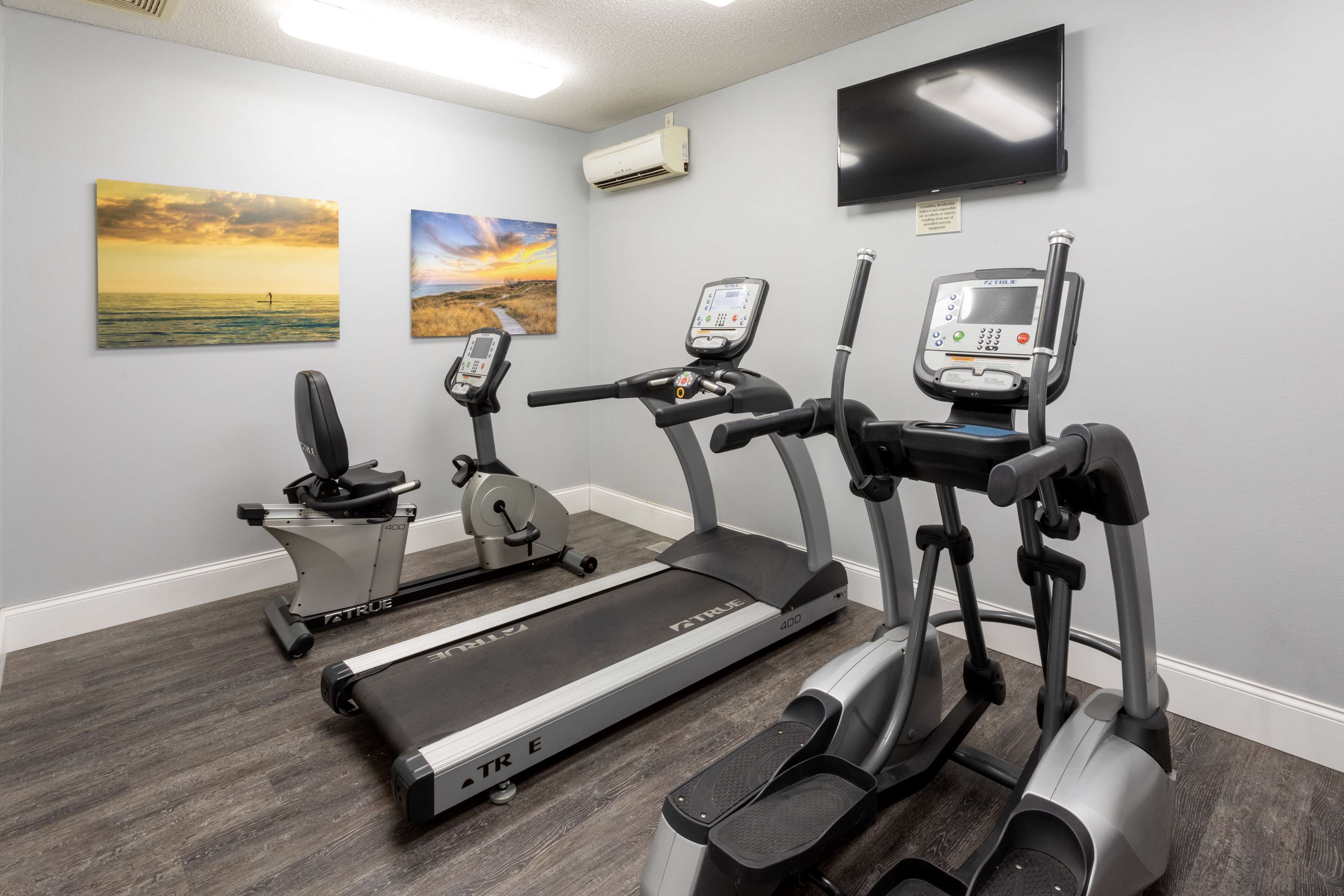 A home gym with treadmills, an elliptical, and a stationary bike, featuring a TV and artwork on the walls.