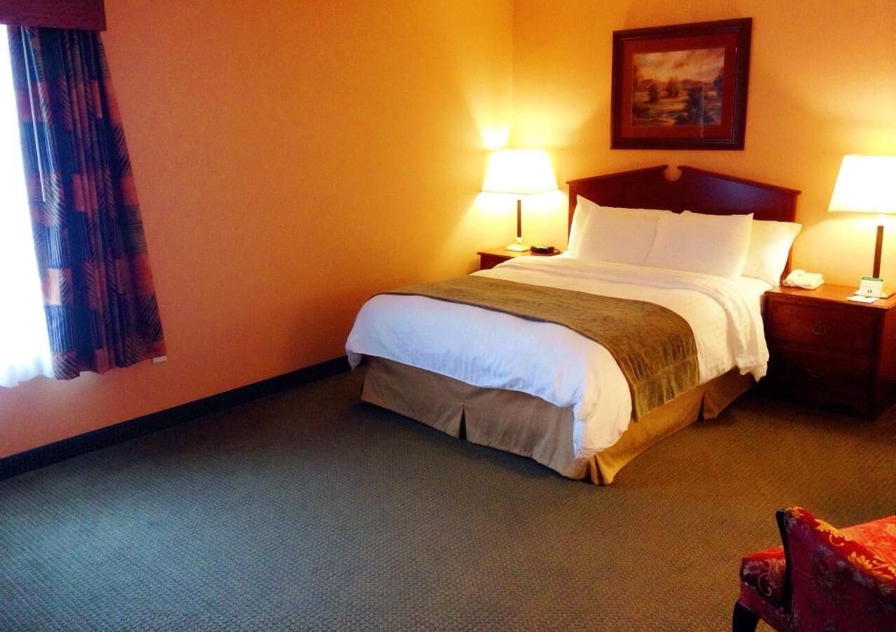 A hotel room with a bed, two bedside tables, a lamp, a chair, and a painting on the wall.